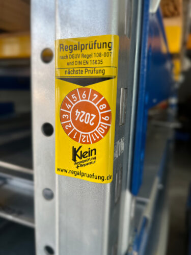 Plakette der bestandenen Regalprüfung, mit Hinweis auf nächste Prüfung in gelber Farbe, aufgeklebt an einem Regal